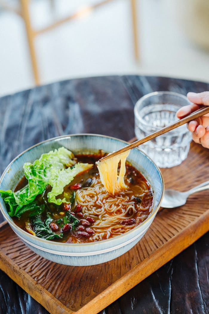 Delicious noodle soup with chopsticks and fresh greens in a ceramic bowl on a wooden tray.
