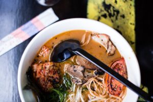 High-angle view of pork ramen with vegetables and sesame seeds in a white bowl.