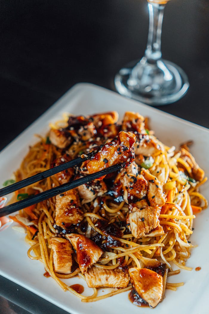 about-us-01 A mouthwatering plate of teriyaki chicken noodles garnished with sauce and served with chopsticks.